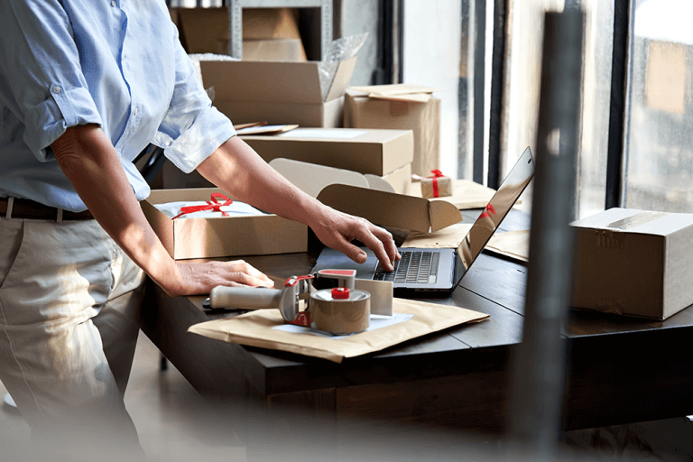 A person wearing a light blue shirt is typing on a laptop keyboard next to a package and tape roller.