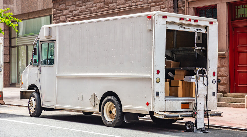 A white delivery van with its back door open, exposing stacked boxes, parked next to a brown brick building