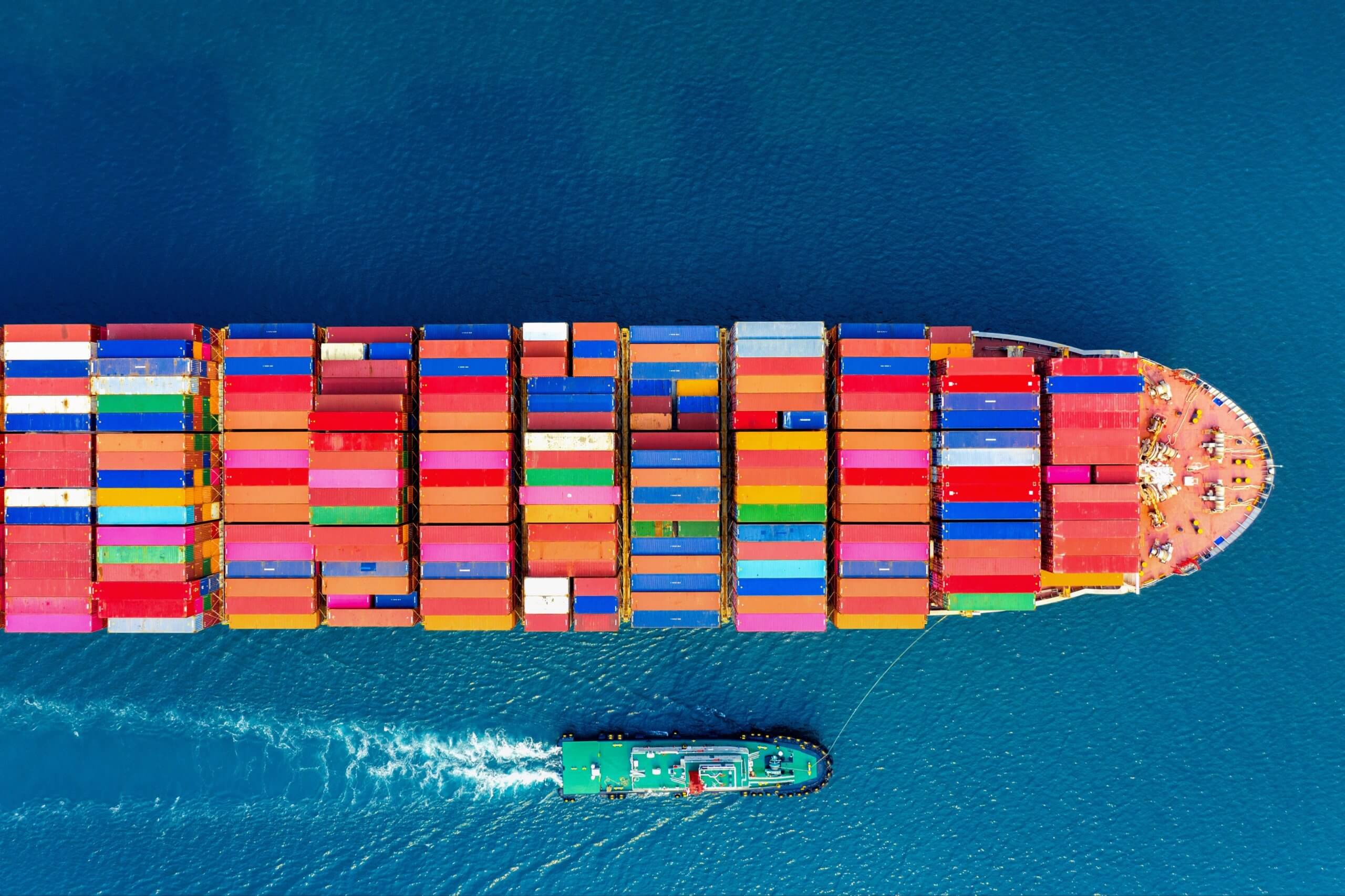 Aerial view of a freight ship carrying colourful cargo containers sails over blue waters, alongside a smaller green pilot ship.