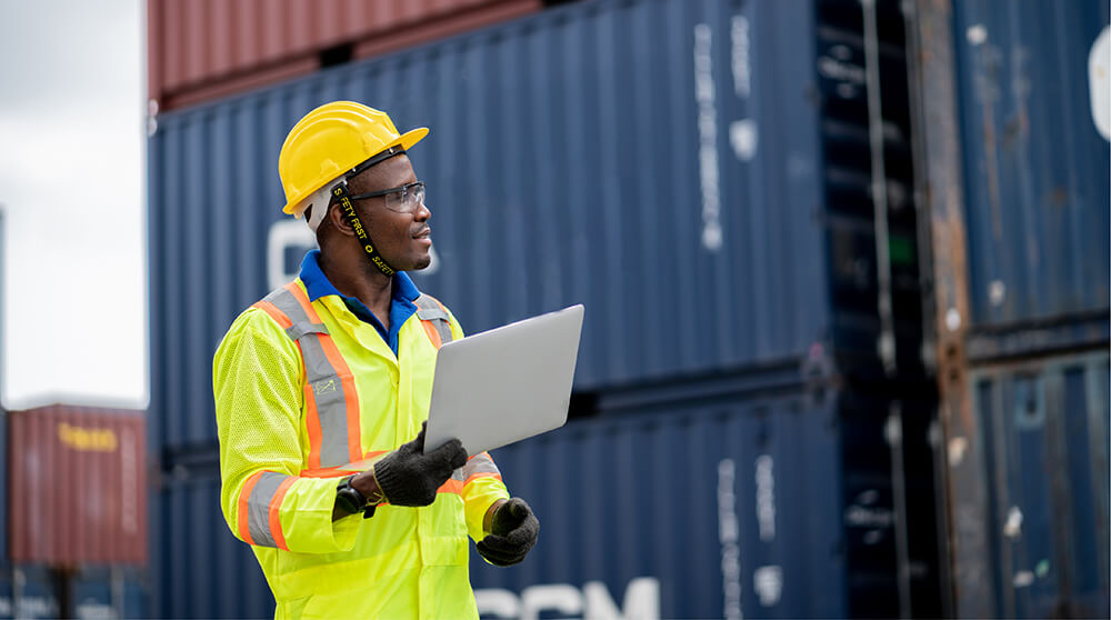 A shipping worker wearing a yellow hardhat and reflective jacket is holding a clipboard and examining cargo containers