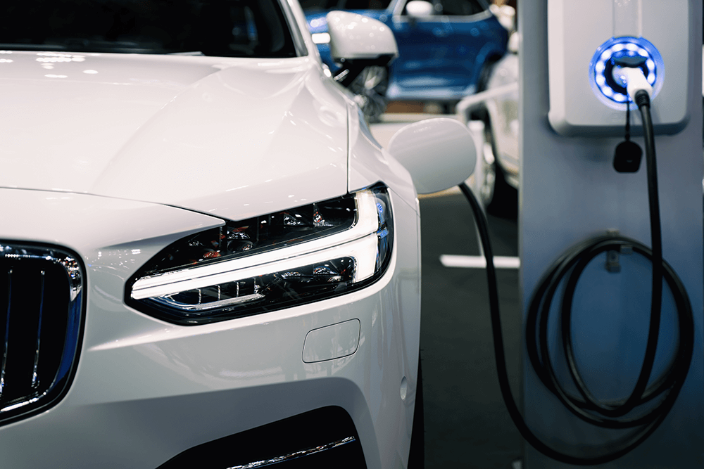 A white electric vehicle is parked at a charging station. A black hose connects the car to a charging port with a neon light.