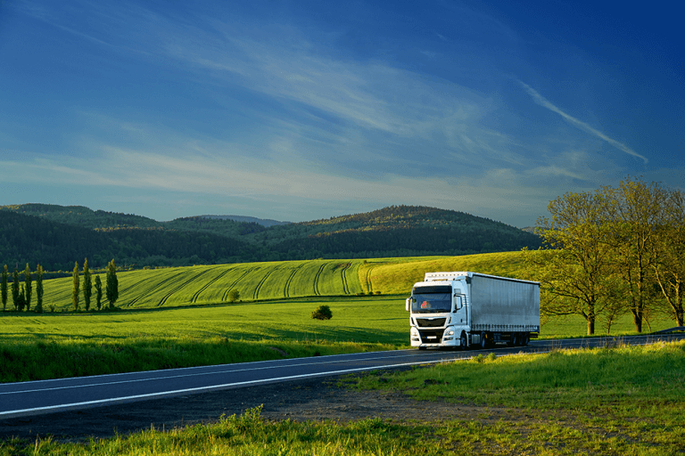 A white transport truck drives on an open highway, surrounded by green fields, forests, hills, and a blue sky