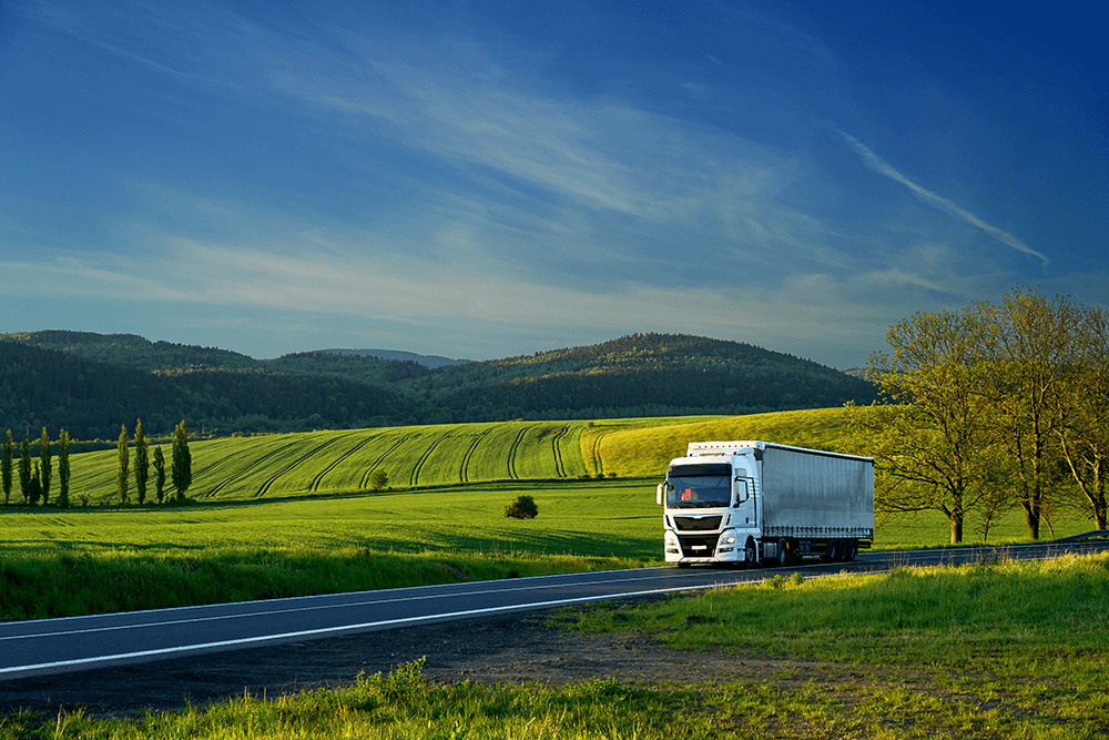 A white transport truck drives on an open highway, surrounded by green fields, forests, hills, and a blue sky