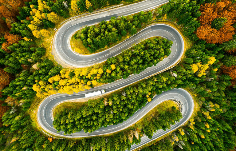A white transport truck is pictured from above driving on a winding road through a colorful autumn forest