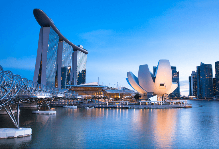 Intricately shaped buildings and bridge reflect off the water in a landscape view Singapore's Marina Bay Sands resort complex in the early evening.