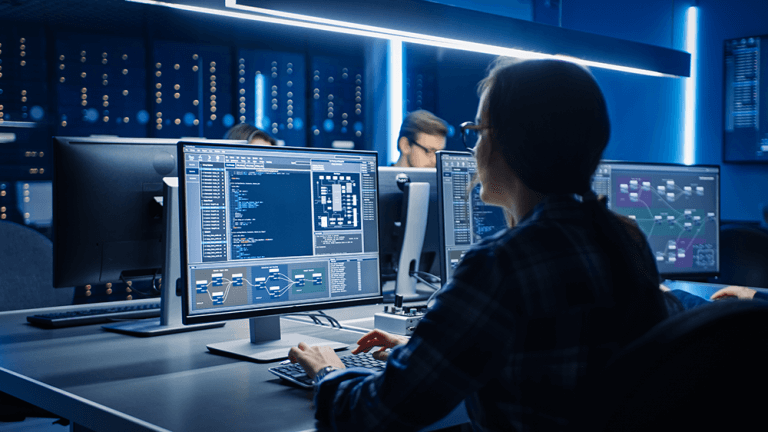 A team of computer engineers sit in front of monitors reviewing code in a dark blue room