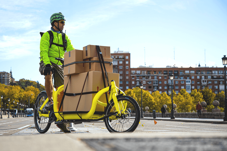 A courier wearing a lime green jacket, shorts, and a watermelon helmet, rides a yellow courier bike hauling several packages in a small city during daytime