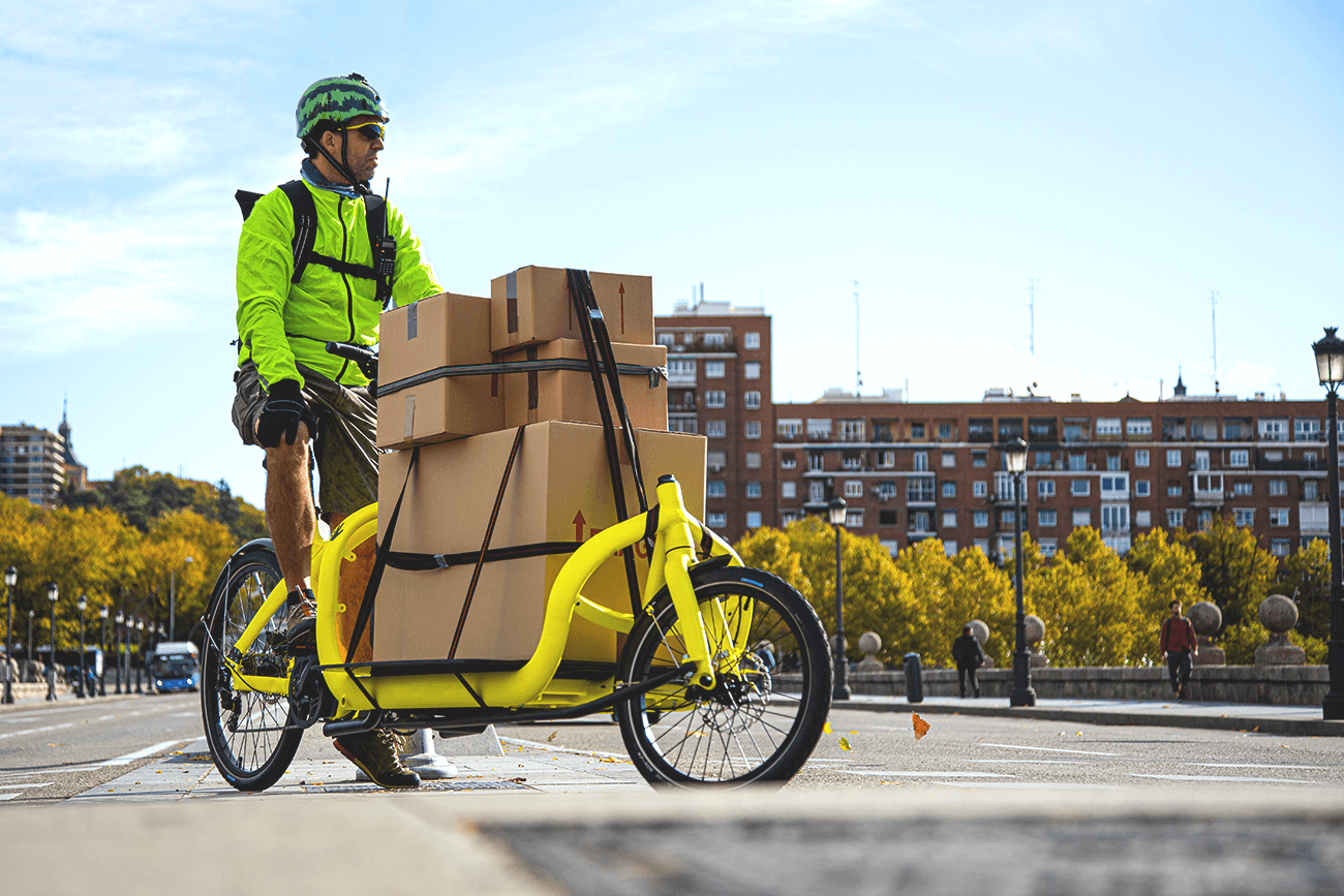 A courier wearing a lime green jacket, shorts, and a watermelon helmet, rides a yellow courier bike hauling several packages in a small city during daytime