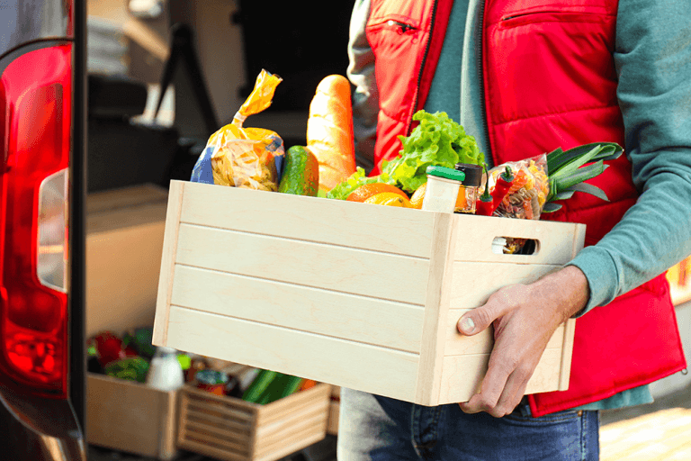 A delivery worker wearing a red vest carries a crate of fresh vegetables at a market.
